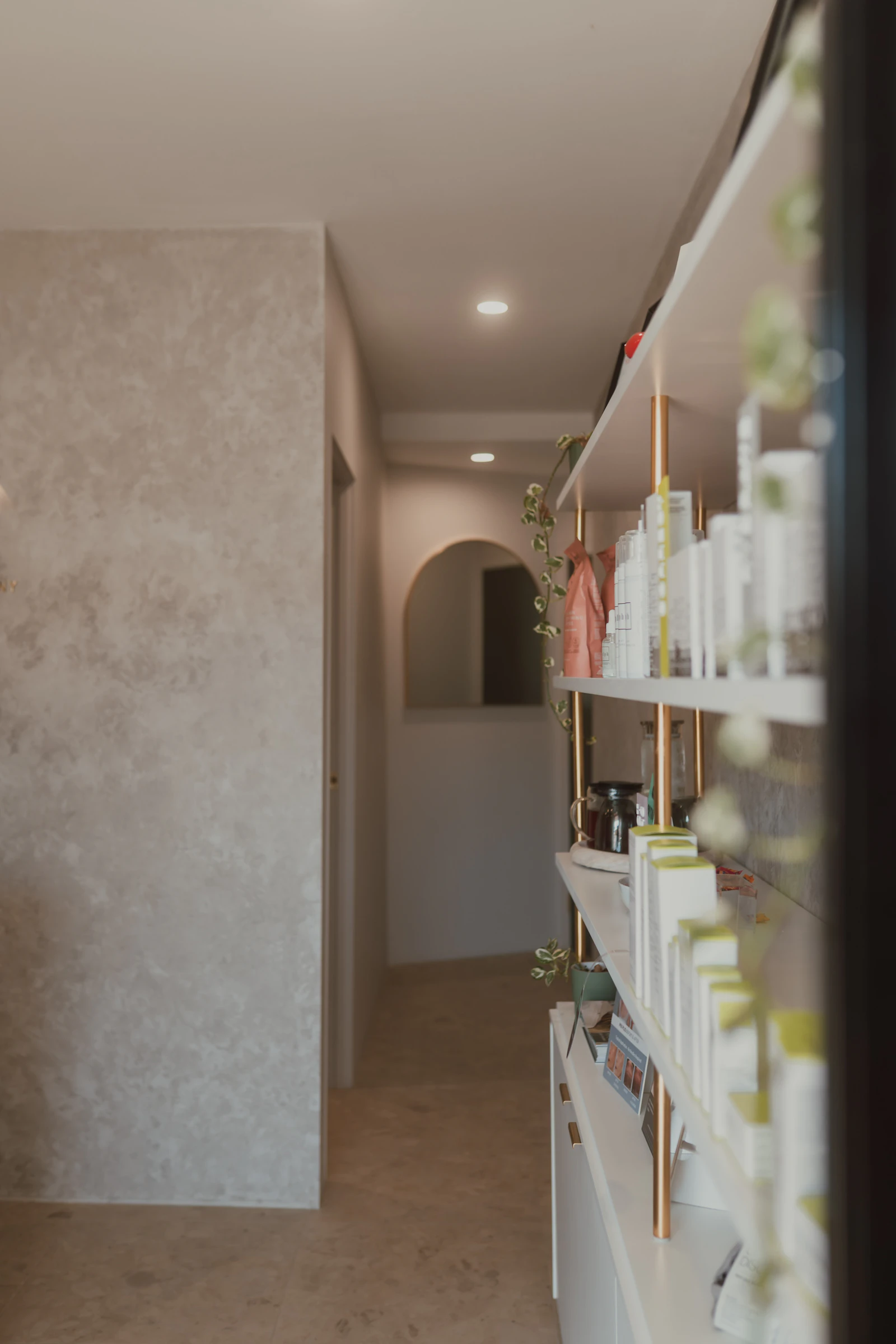 A corridor in the clinic with white shelves of skincare on copper uprights and an arched mirror at the far end